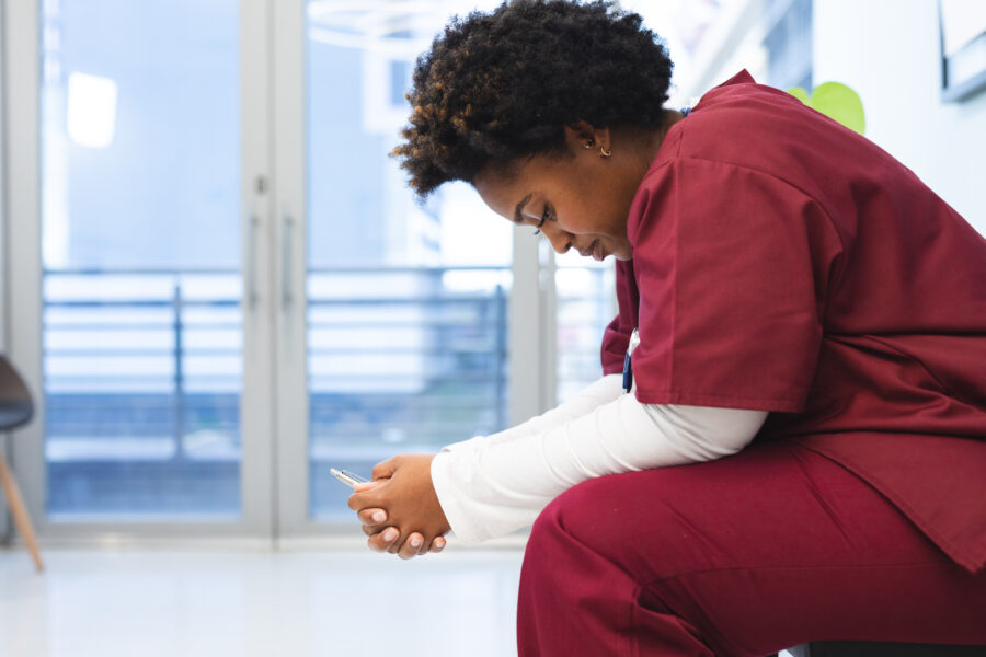 Side view of a nurse in scrubs sitting with her head down, leaning her arms on her knees and hands clasped. She looks tired. A hospital wall or door is in the background. 