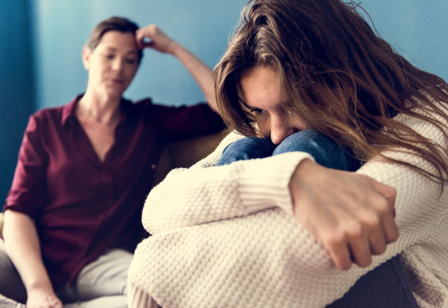 In the foreground a teen sits hugging their knees and looking looking distressed, they are wearing a white sweater and jeans. In the background just slightly out of focus a parental figure sits and looks stressed with their head leading a bit into their hand. They are sitting on the same couch. 